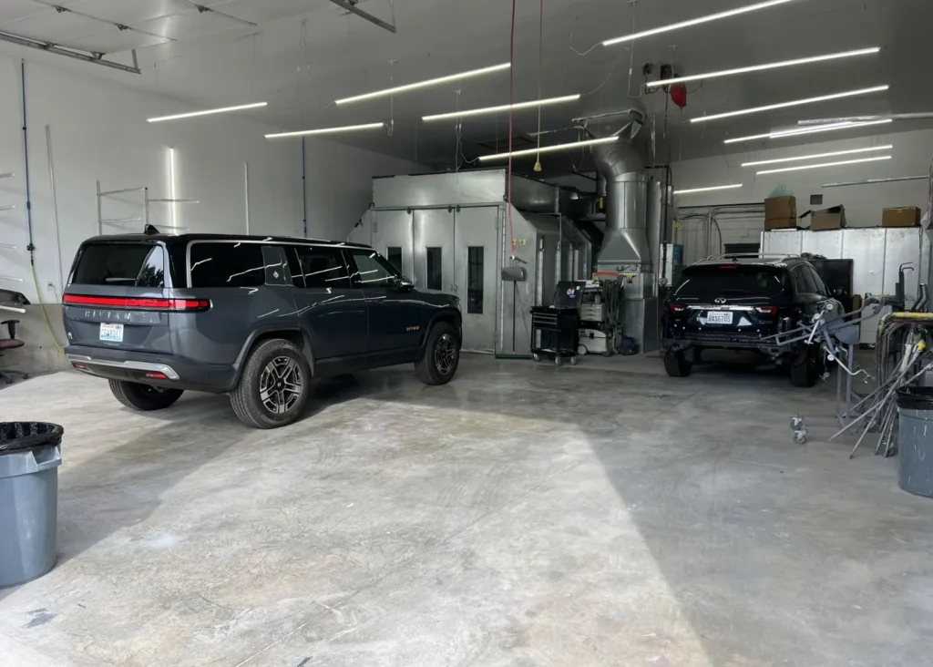 Dark gray Rivian R1S electric SUV and Infiniti undergoing service inside an auto body shop paint booth bay.